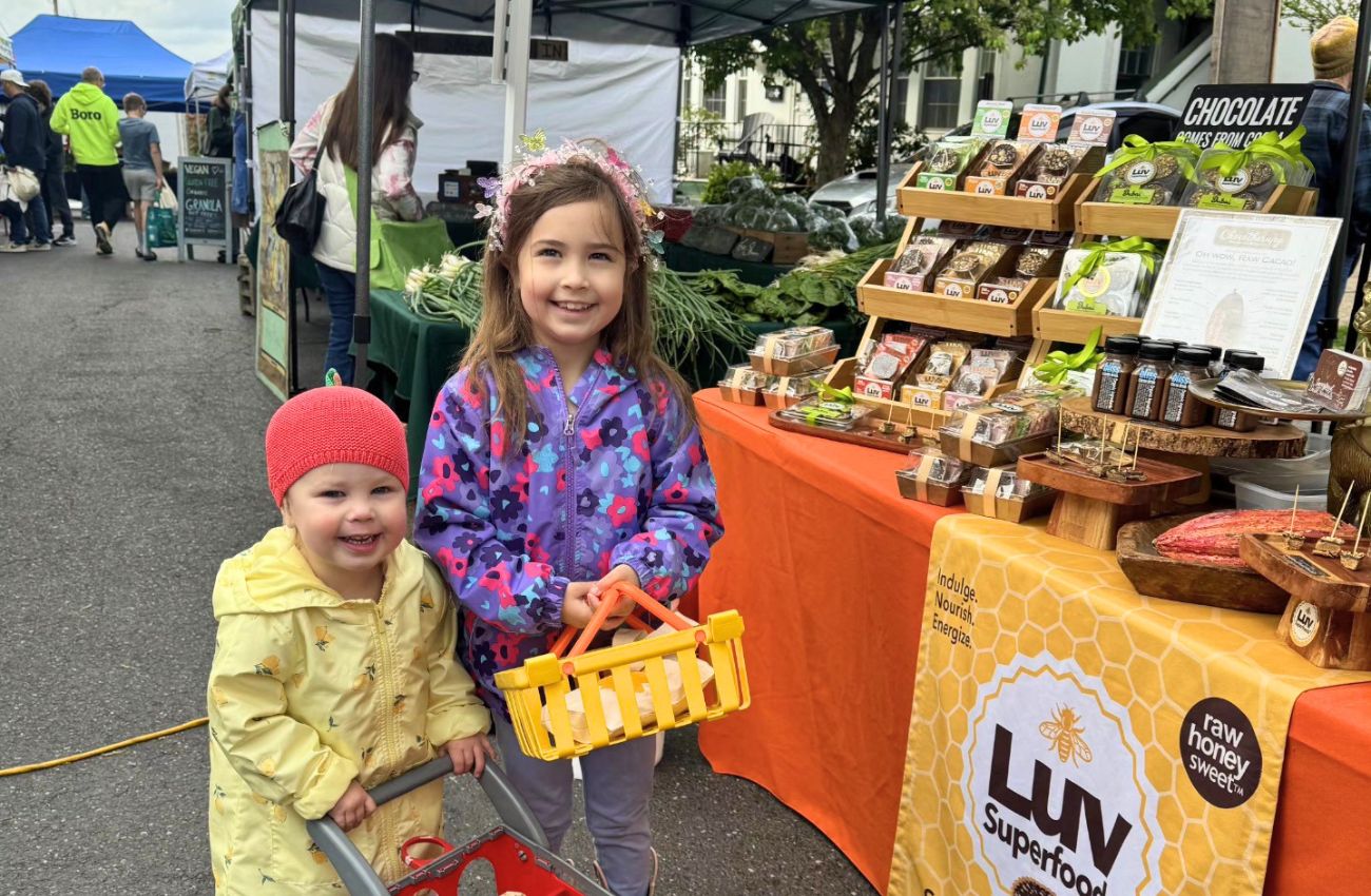Two little girls hold a shopping basket and cart at the Doylestown Farmers Market.