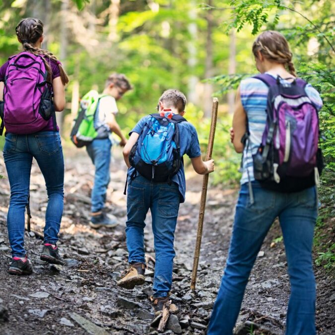 A mom and three kids wear backpacks on a woods hike.