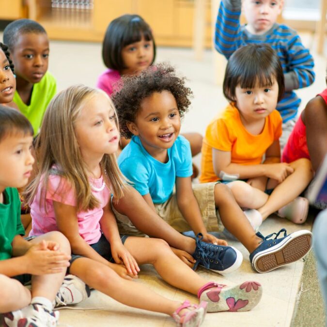 A group of kindergarten age children sit on the floor listening to an adult read a story.