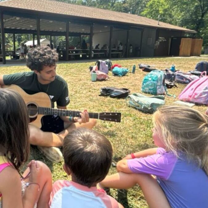 A young adult camp counselor plays guitar for a group of young summer camp kids sitting on the grass.