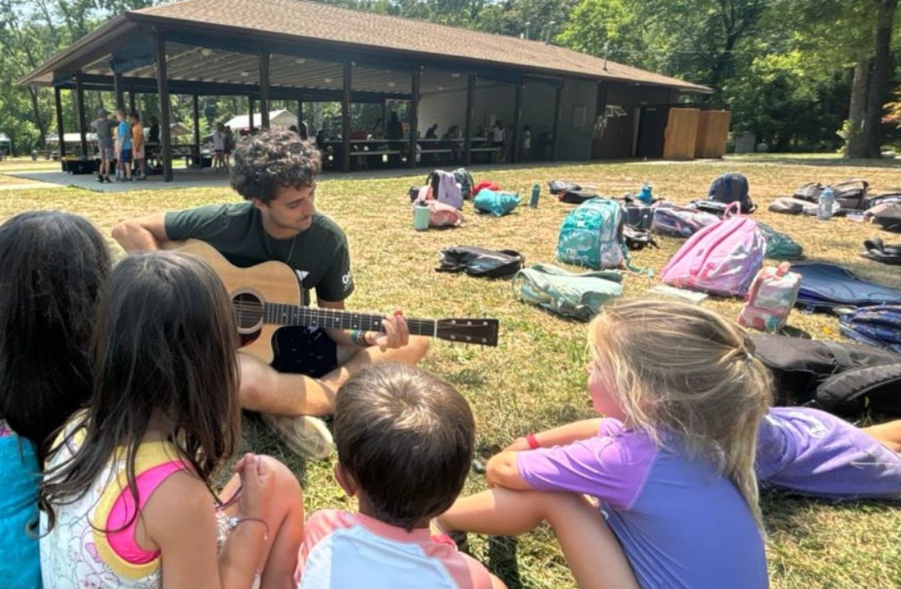 A young adult camp counselor plays guitar for a group of young summer camp kids sitting on the grass.