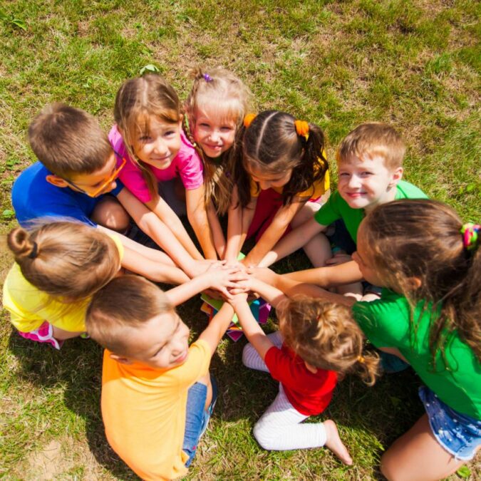 A group of preschool kids sit in a circle on the grass at summer camp.