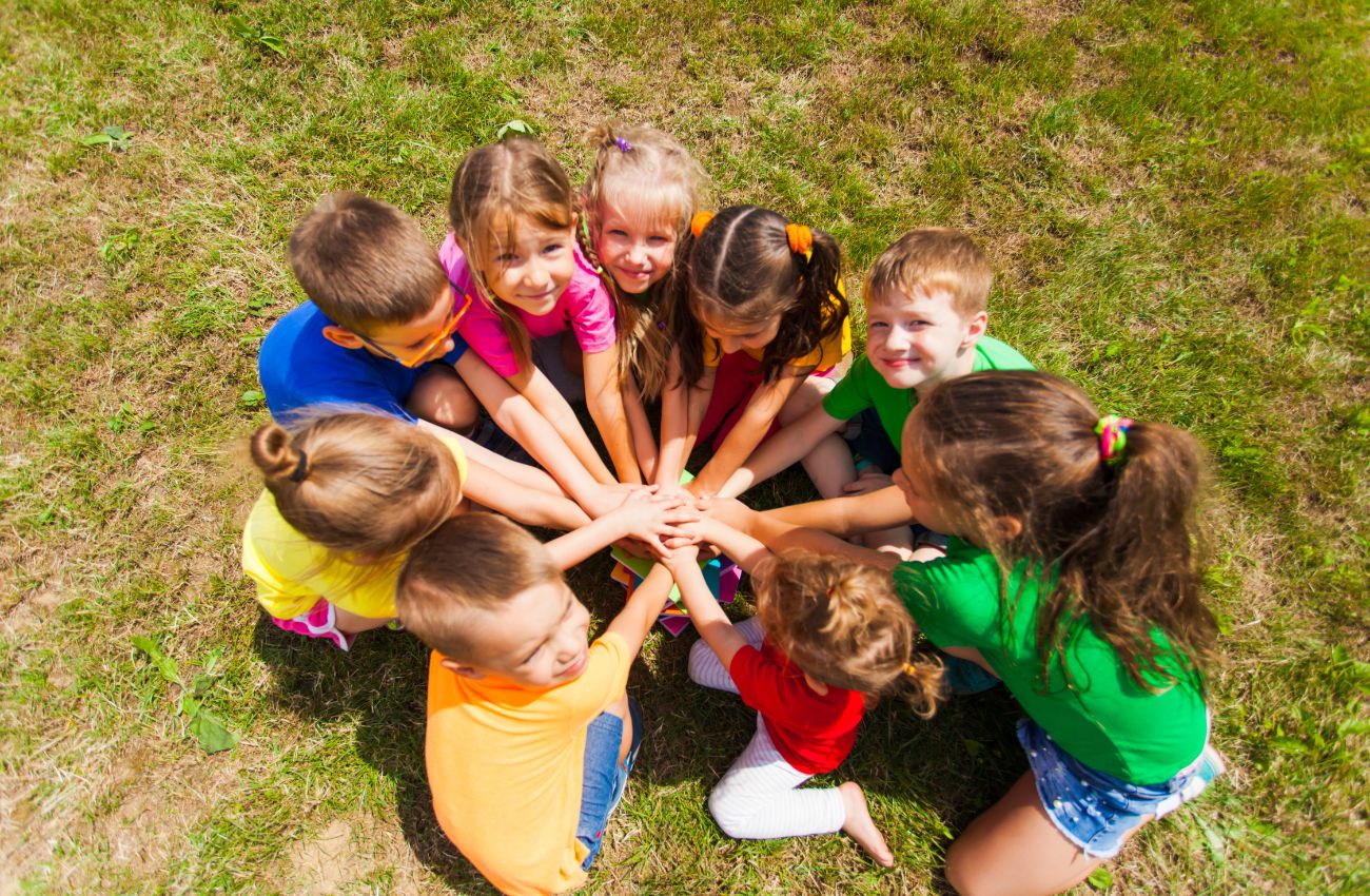 A group of preschool kids sit in a circle on the grass at summer camp.