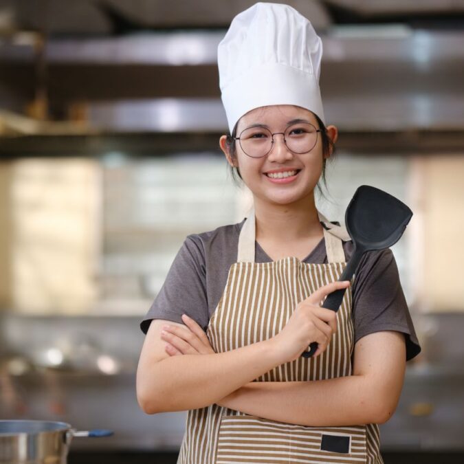 A teen girl stands in a kitchen wearing a chef hat and holding a spatula.
