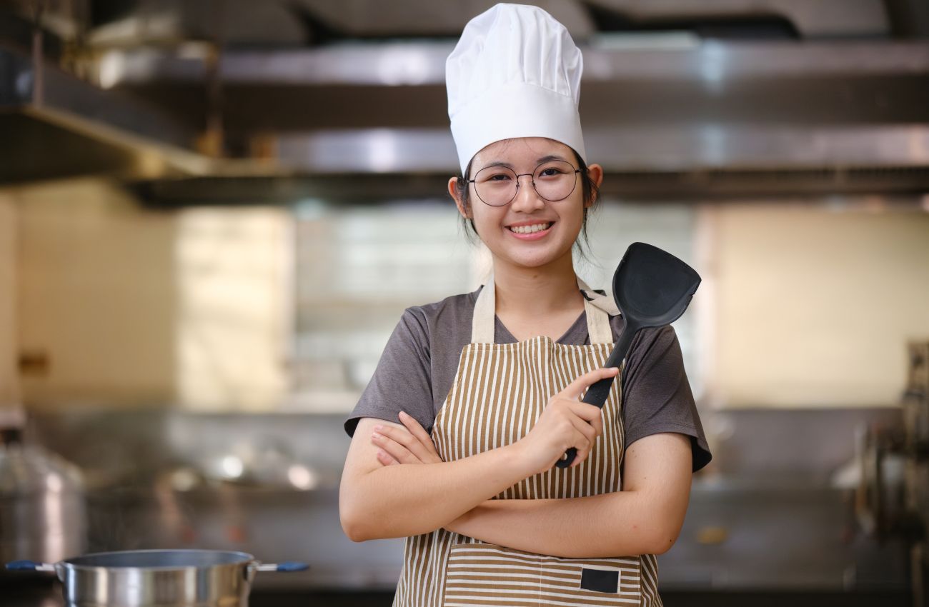 A teen girl stands in a kitchen wearing a chef hat and holding a spatula.